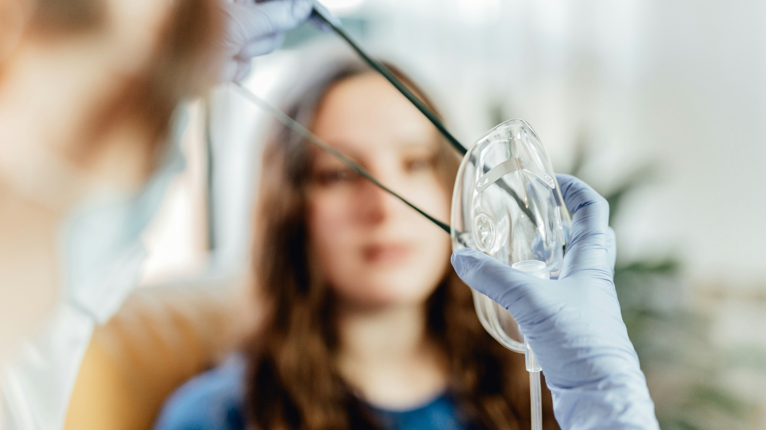 Gloved hands hold out an oxygen mask, a blurred woman sits in the background.